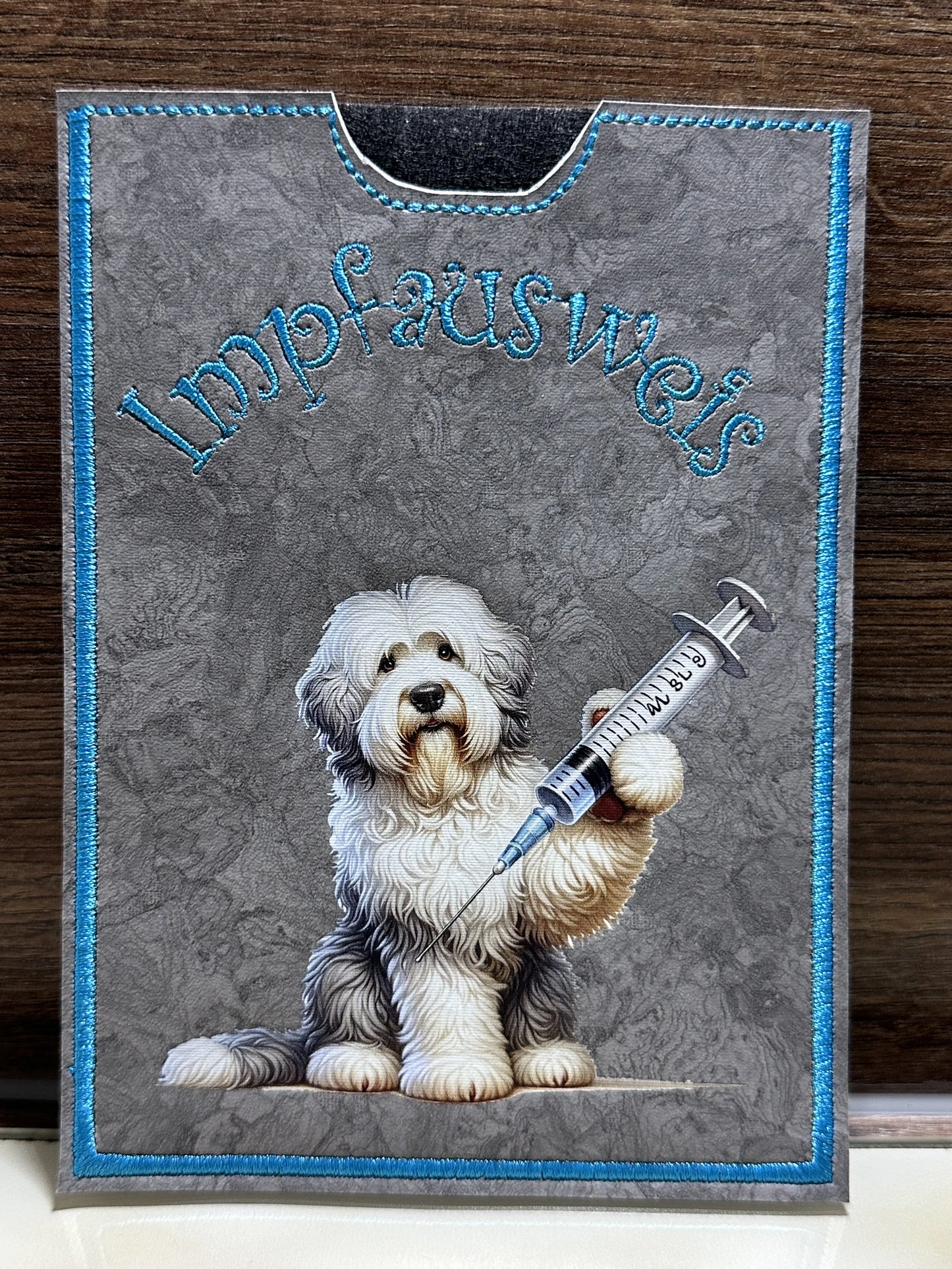Handgemachte Impfpasshülle 12,5 × 17 cm aus grauem Kunstleder mit blauem Rand und Bobtail-Motiv mit Spritze, passend für EU-Heimtierausweis